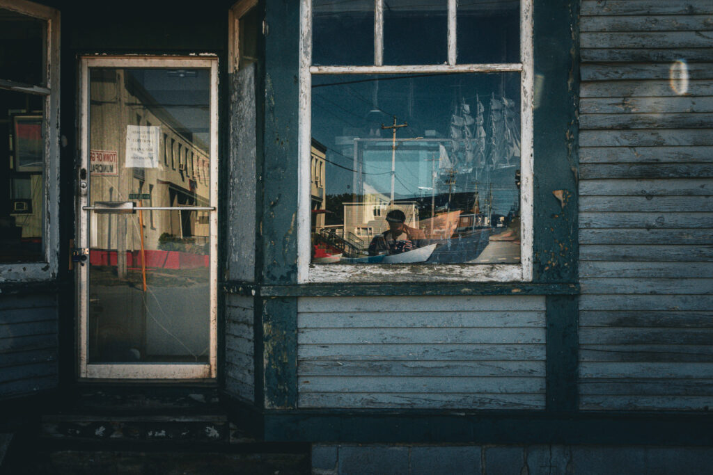 A blue storefront with a large window and glass door. The exterior looks somewhat neglected, there is a model ship in the window, the photographer's reflection, and the composition is moody.